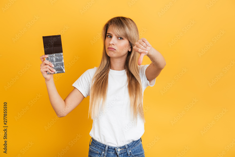 Young blonde woman posing isolated over yellow wall background dressed in white casual t-shirt holding chocolate sweeties showing thumbs down. Young blonde woman posing isolated over yellow wall background dressed in white casual t-shirt holding chocolate sweeties showing thumbs down.