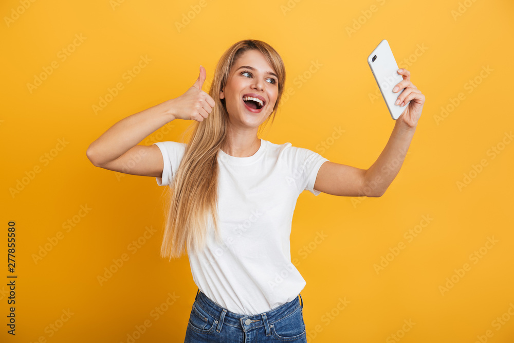 Happy young blonde woman posing isolated over yellow wall background dressed in white casual t-shirt using mobile phone take a selfie. Happy young blonde woman posing isolated over yellow wall background dressed in white casual t-shirt using mobile phone take a selfie.