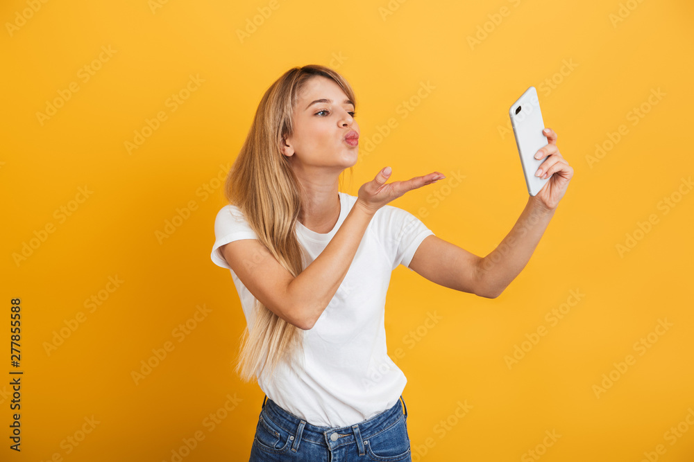 Happy young blonde woman posing isolated over yellow wall background dressed in white casual t-shirt using mobile phone take a selfie. Happy young blonde woman posing isolated over yellow wall background dressed in white casual t-shirt using mobile phone take a selfie.