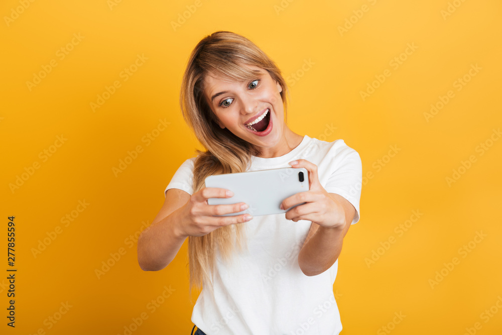 Happy emotional young blonde woman posing isolated over yellow wall background dressed in white casual t-shirt using mobile phone play games. Happy emotional young blonde woman posing isolated over yellow wall background dressed in white casual t-shirt using mobile phone play games.
