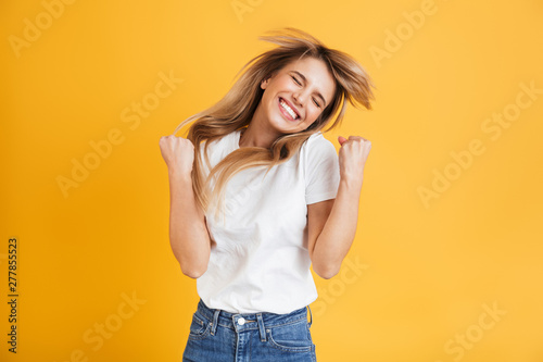 Emotional young blonde woman posing isolated over yellow wall background dressed in white casual t-shirt showing winner gesture. photo