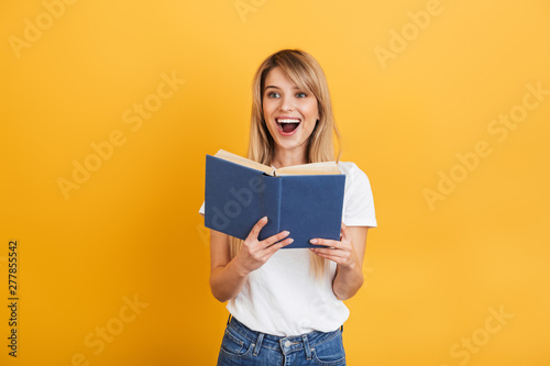 Cheerful emotional young blonde woman posing isolated over yellow wall background dressed in white casual t-shirt holding book reading.