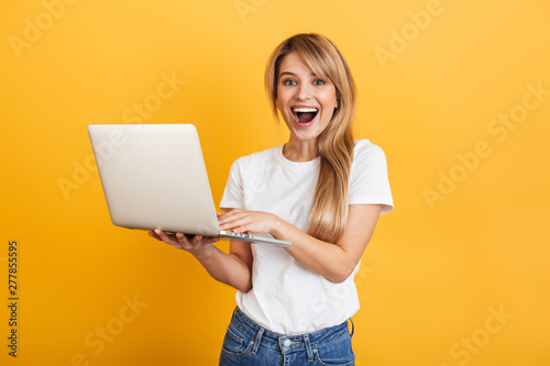 Positive optimistic emotional young blonde woman posing isolated over yellow wall background dressed in white casual t-shirt using laptop computer.