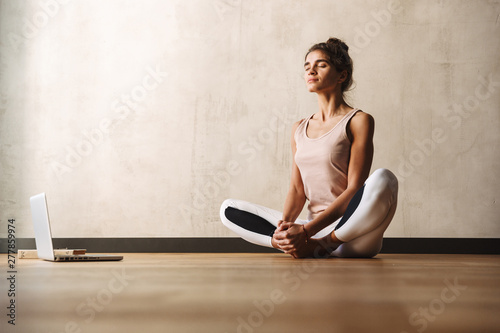 Photo of concentrated nice woman doing exercises with closed eyes and using laptop while sitting on floor at home