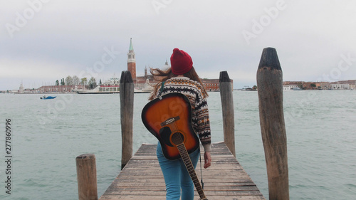 A young attractive woman walks along the pier with a guitar behind her back.