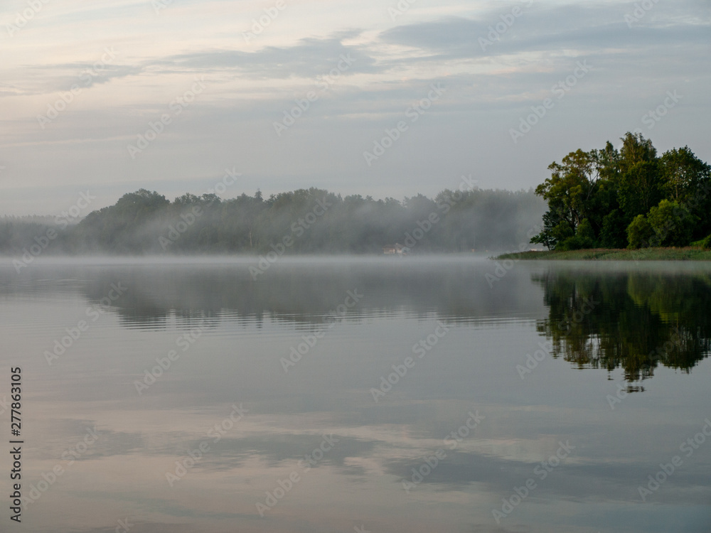 Fototapeta premium Foggy and mystical lake landscape before sunrise. All silhouettes are blurry and unclear. Vaidavas lake, Latvia