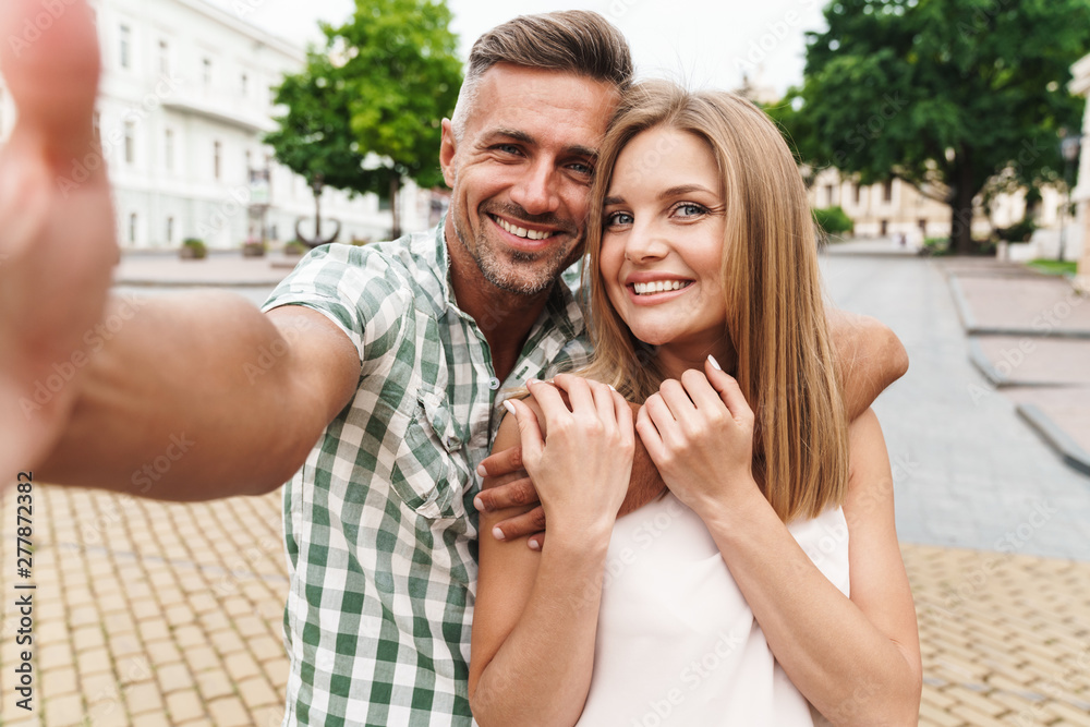 Image of charming young couple smiling and taking selfie photo together while walking through city street