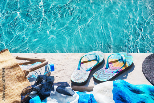 Swimming pool and straw hat, colorful flip flops, wicker bag, straw hat, bottle with still water as sunny summer background. Travel holiday suntan vacation concept. Flat lay, top view, close up.