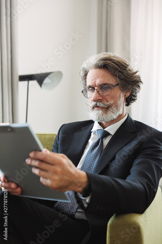Portrait closeup of successful mature businessman holding clipboard while sitting on armchair in apartment