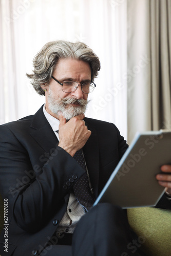 Portrait closeup of working mature businessman holding clipboard while sitting on armchair in apartment
