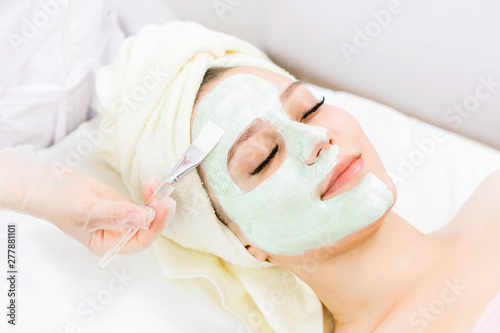 Portrait of beautiful woman laying with towel on the head. Young girl enjoys green cream facial mask. Lady getting spa treatment at beauty salon.