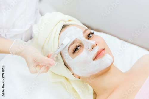 Portrait of beautiful woman laying with towel on the head. Hands of cosmetology specialist applying cream facial mask using brush. Model with brown eyes and healthy glow perfect smooth skin.