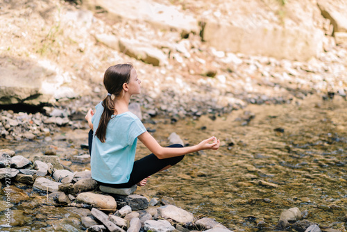 Young girl yoga meditating at sunrise sitting on stones on the shore of a mountain stream. Teenage model meditating in peaceful harmony