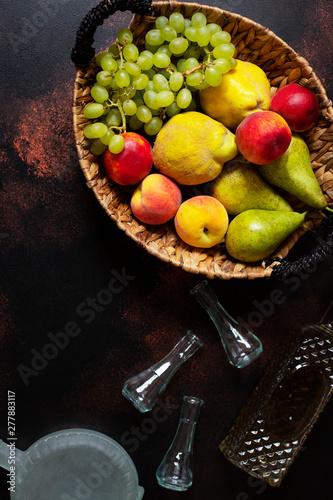 Rakia or Rakija the traditional balkan fruit brandy. The fruits of which produce Rakia. Fruits in a basket, bottles and glasses for alcohol. Top view. Dark vintage background. Space for text photo