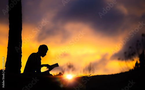 Silhouette of young male christian sitting and reading the holy Bible with light of sunset background, christian hope concept.