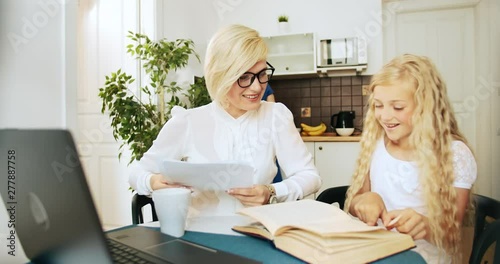 Young pretty business lady hugging cute blond daughter, sitting near and reading book, family bonds