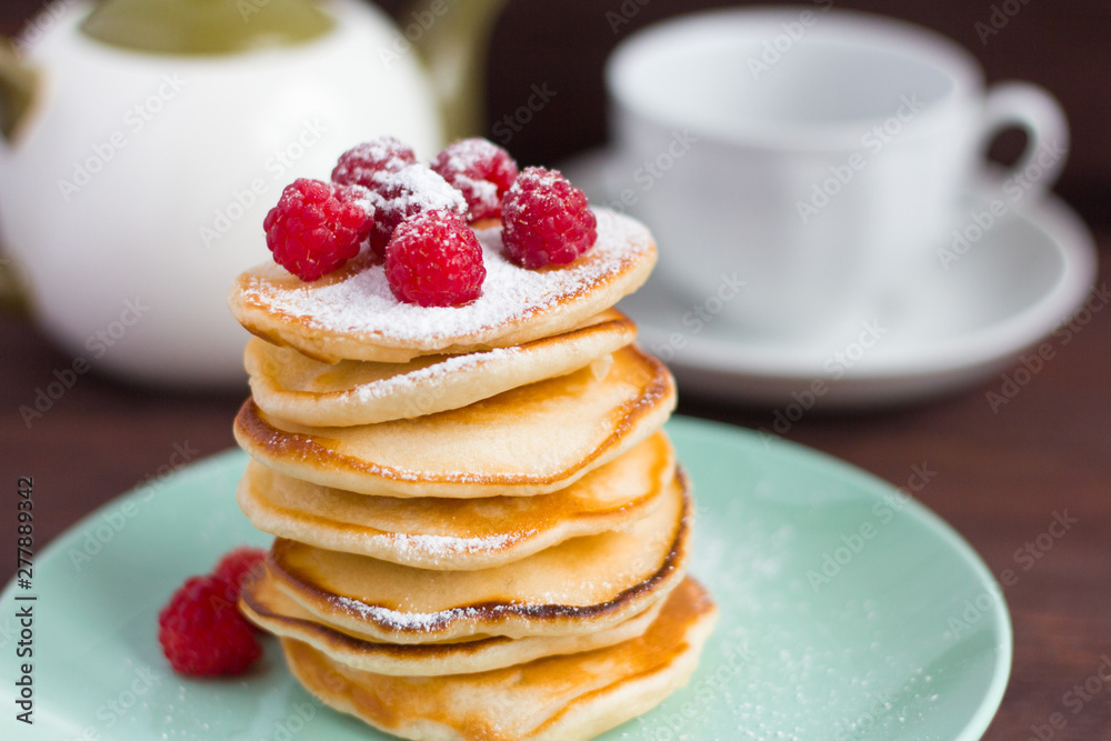 stack of baked homemade pancakes with raspberries, sprinkled with powdered sugar and tea utensils in the background, selective focus