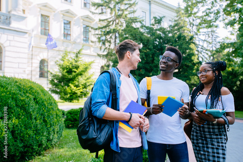 Portrait of three happy high school multiethnic students from different countries walking to class, after lesson, outdoors holding books in european city. Multinational friendship concept.