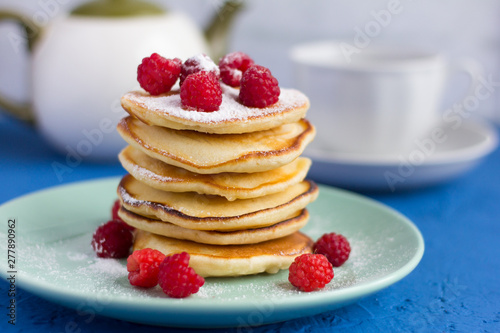 stack of baked homemade pancakes with raspberries  sprinkled with powdered sugar and tea utensils in the background  selective focus