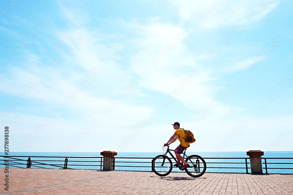 Obraz premium Young man with fit body riding the mtb mountain bike on sandy beach with beautiful azure water sea view. Muscle male wearing bright yellow t-shirt cycling on ocean shore. Close up, copy space.