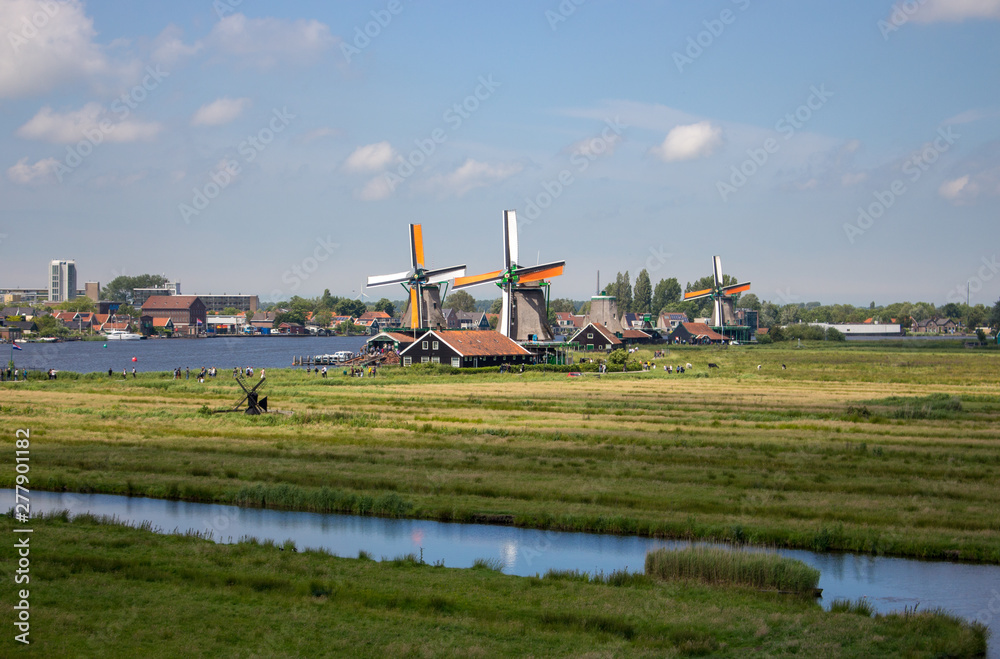 Fototapeta premium Zaanse Schans, Netherlands - 06/14/2019: Old dutch windmills and river in historical village. Holland mills in field panoramic view. Rural holland landscape. Wind energy concept. 