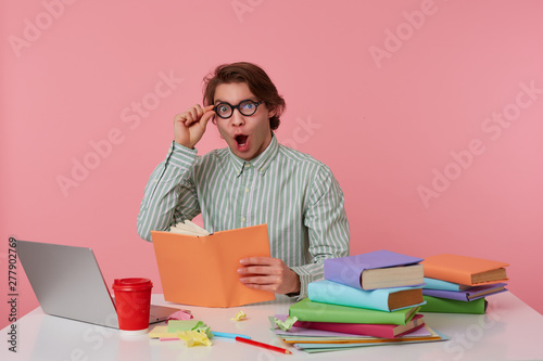 Young wondered student in glasses wears in basic shirt, man sits by the table and working with laptop, looks at the camera through glasses with shocked expression, isolated over pink background. photo