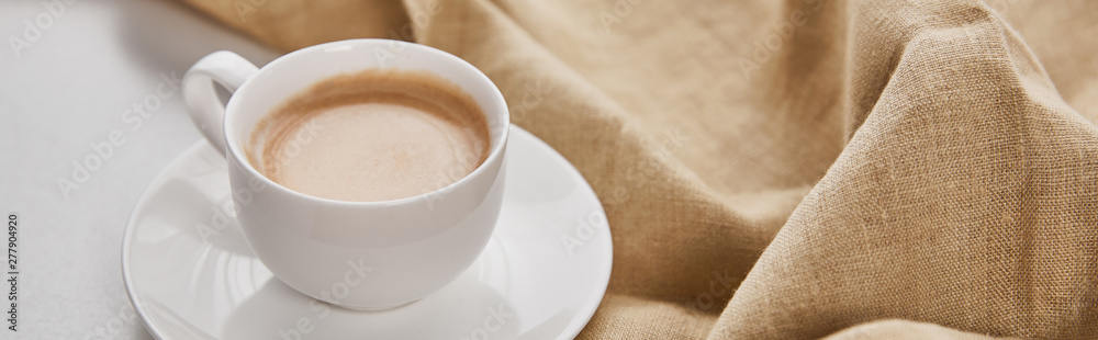 close up view of coffee in white cup on saucer near beige napkin, panoramic shot close up view of coffee in white cup on saucer near beige napkin, panoramic shot