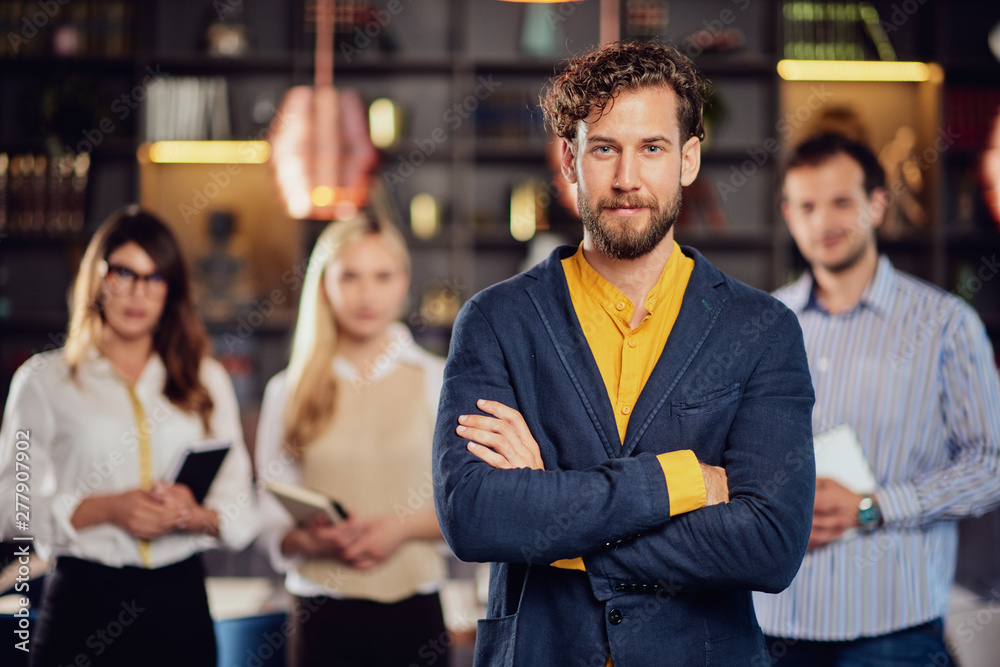 Serious Caucasian businessman dressed smart causal standing in restaurant with arms crossed. In background his successful team posing. Serious Caucasian businessman dressed smart causal standing in restaurant with arms crossed. In background his successful team posing.