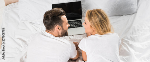panoramic shot of couple lying in bed and using laptop at home