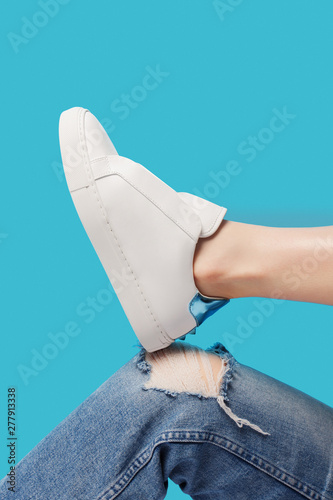 Trendy white eco leather sheakers on bent leg knee in ragged jeans over pastel turquoise blue background. Colorful main trend backdrop for your magazine, online store and media. Copy space
