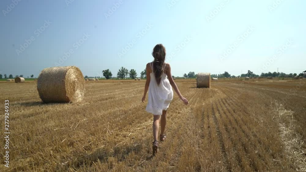 Young woman in white dress goes in golden mown wheat field among hay bales. Agricultural golden field landscape. Young woman in white dress goes in golden mown wheat field among hay bales. Agricultural golden field landscape.