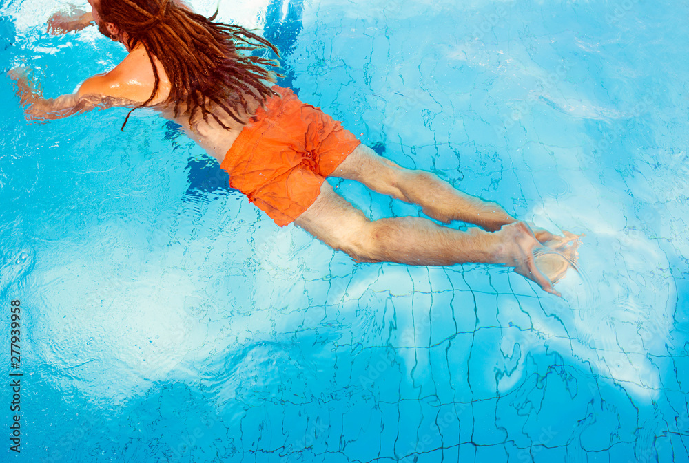 caucasian man back with dreadlocks in orange shorts swimming in the water in the swimming pool in sunny day. Vacation and sport concept.