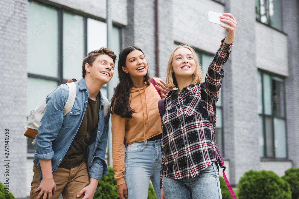 happy teenagers holding smartphone, taking selfie and smiling outside