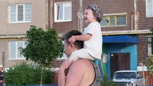 A toddler sits on his father's shoulders and laughs in the yard of a high-rise building