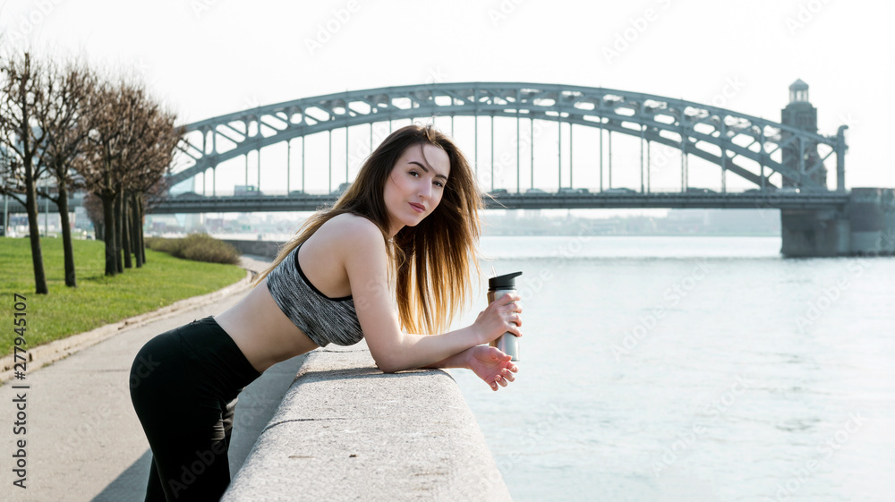beautiful girl with long hair in sportswear with a bottle stands on the Neva river embankment against the bridge