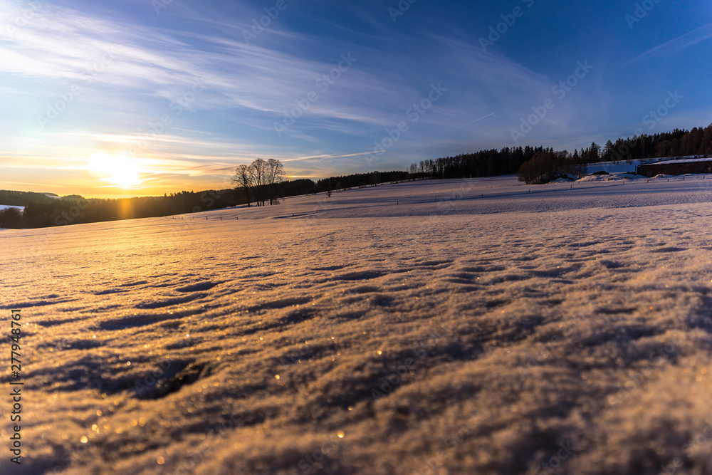 Beautiful winter sunset over a hilly landscape. Vast meadow covered in snow. Sun peaking over the horizont, orange and yellow colors, blue sky. 