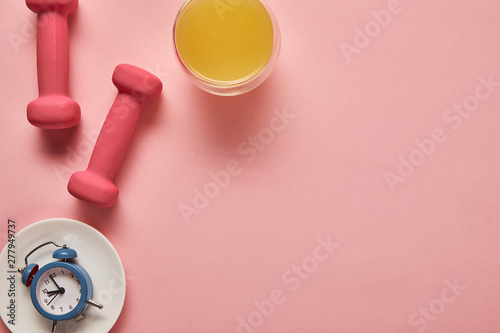 top view of orange juice, pink dumbbells and toy alarm clock on plate on pink background