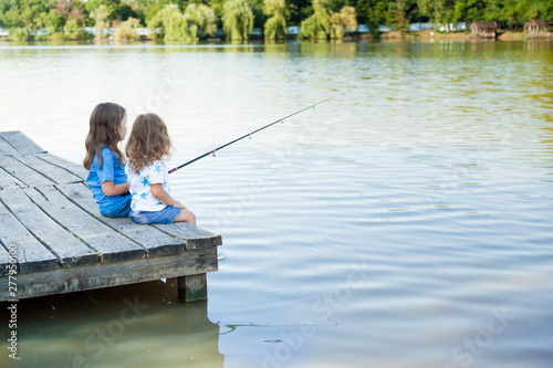 children are fishing from wooden pier on a lake. two cute sisters have fun fishing. Family leisure activity during summer sunny day. Active family time on nature. Hiking and adventure with little kid 