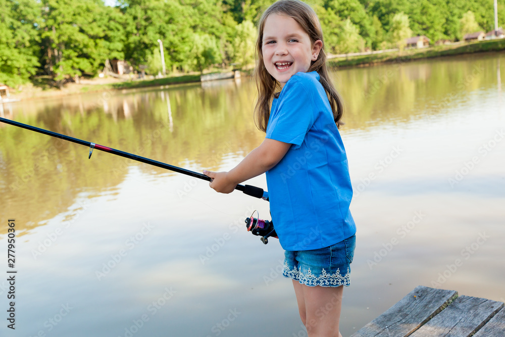 Cute child girl fishing from wooden pier on a lake. Family leisure activity during summer sunny day. little girl having fun by a river at beautiful summer evening Cute child girl fishing from wooden pier on a lake. Family leisure activity during summer sunny day. little girl having fun by a river at beautiful summer evening