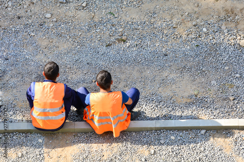 Migrants workers in yellow and orange vests resting by the road. They are sitting on the sidelines. Repair the road. Copy space. The concept of a break for rest, lack of work, unemployment.