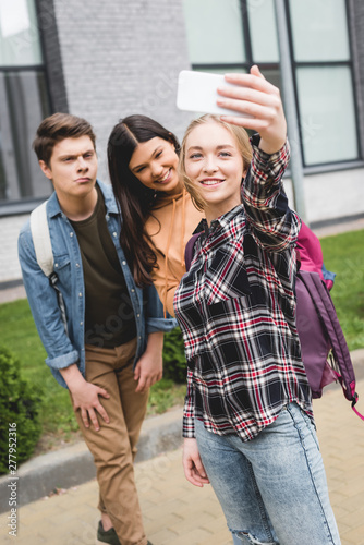 positive teenagers holding smartphone  taking selfie and smiling outside
