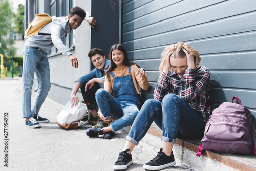 smiling friends bulling and pointing with fingers at sad blonde teen