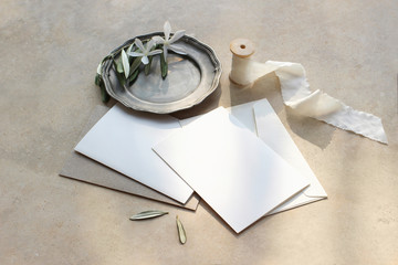 Summer wedding stationery mock-up scene. Blank greeting cards, envelopes, silver plate with olive branch and white flowers and silk ribbon. Marble background in sunlight, shadows. Top view.