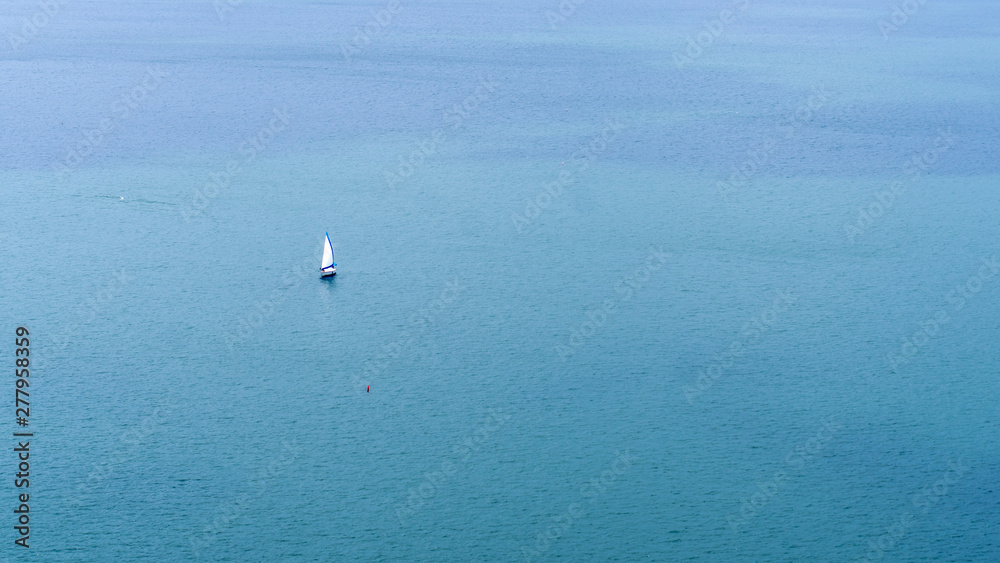 Small white and blue sailboat on calm, open blue water with small red buoy nearby