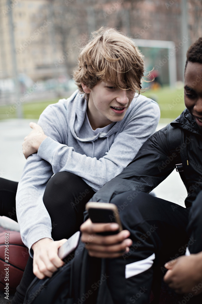 Teenage boy looking at friend's mobile phone while sitting on sidewalk after basketball practice Teenage boy looking at friend's mobile phone while sitting on sidewalk after basketball practice