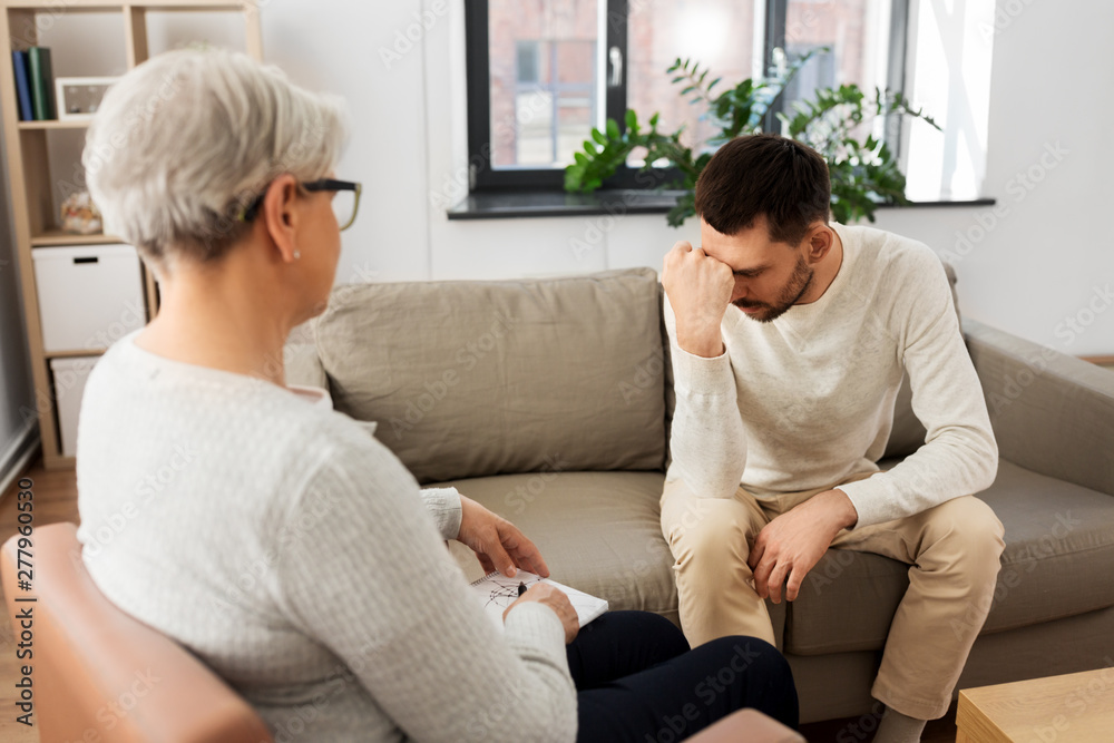 psychology, mental therapy and problem concept - senior woman psychologist taking notes to notebook and sad young man patient at psychotherapy session