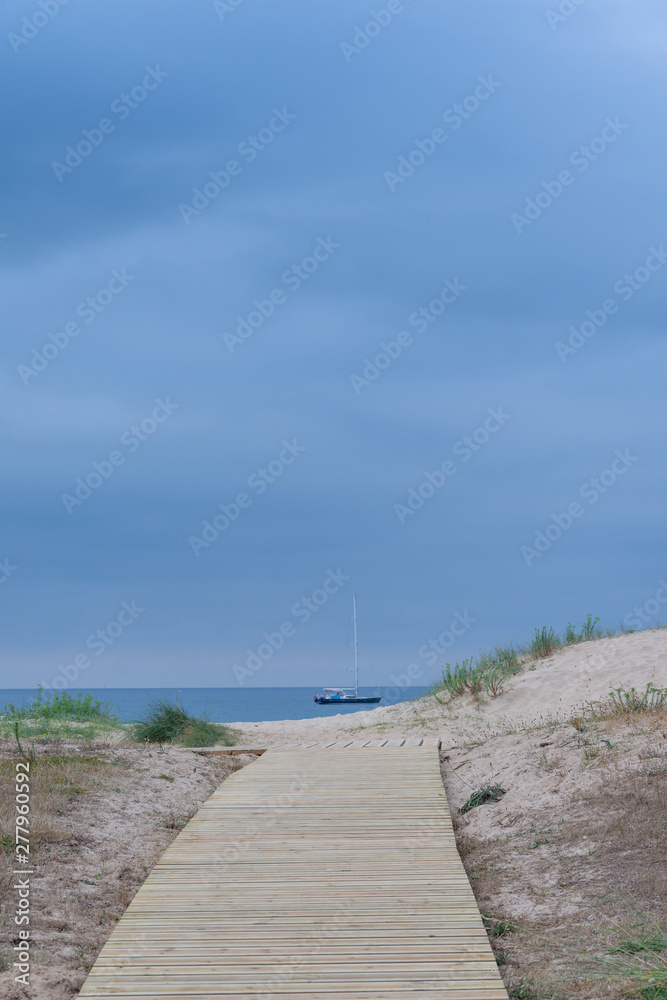 Wooden path to the beach with yacht in the sea and cloudy sky in the background in Cantabria, Spain, Europe