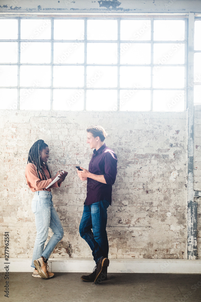 Full length side view of young IT professionals talking while standing by weathered wall in office Full length side view of young IT professionals talking while standing by weathered wall in office