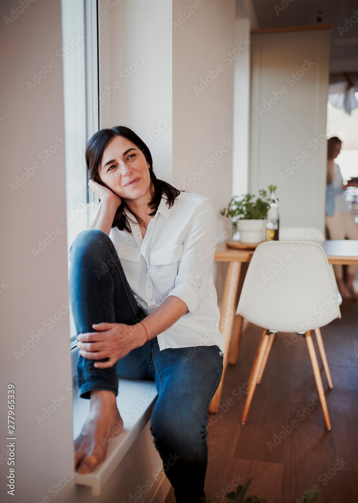 Mature woman sitting on windowsill in the kitchen, daydreaming Mature woman sitting on windowsill in the kitchen, daydreaming