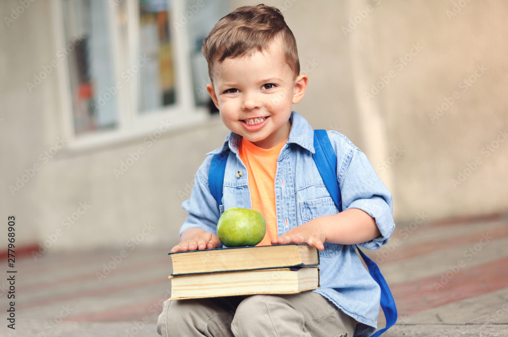 Fototapeta premium A small three year old preschooler with a backpack on his back and with books sits on the stairs and shows his green apple for a snack.
